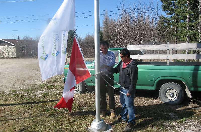 Two individuals are raising flags on a pole in an outdoor setting, with a green pickup truck visible in the background. The scene captures a moment of flag-raising, likely during a ceremony.