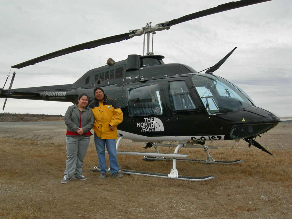 Two people stand next to a black helicopter on a grassy area under a cloudy sky. The individuals are casually dressed, with one wearing a bright yellow jacket.