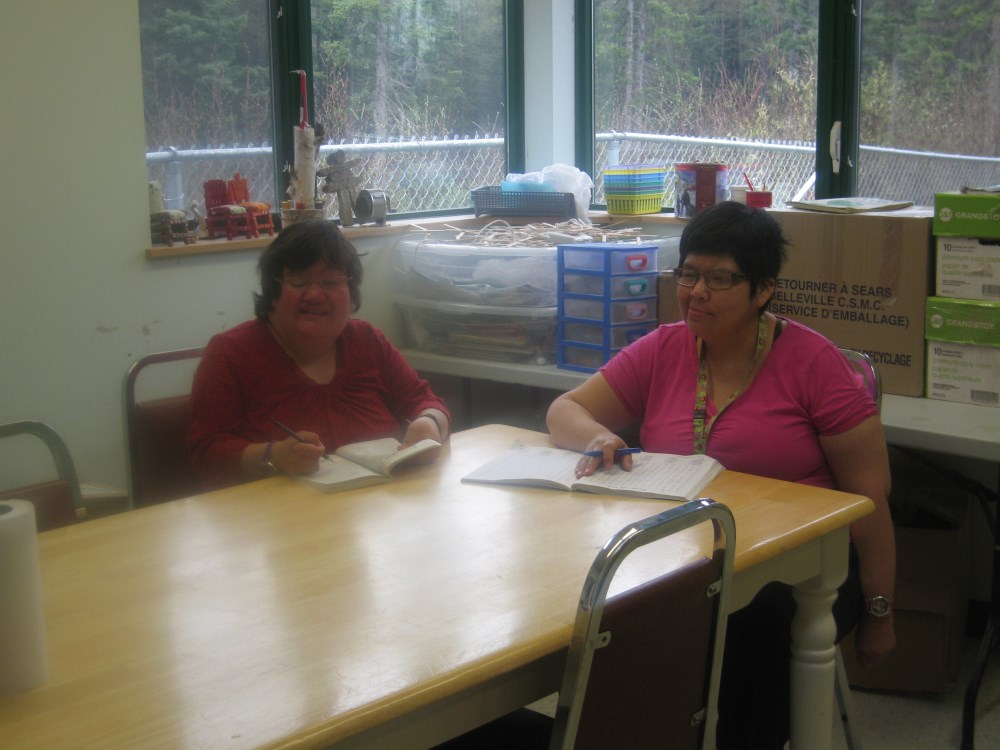 Two women are seated at a table in a bright room, writing in notebooks. Boxes are stacked in the background, suggesting an organized workspace.
