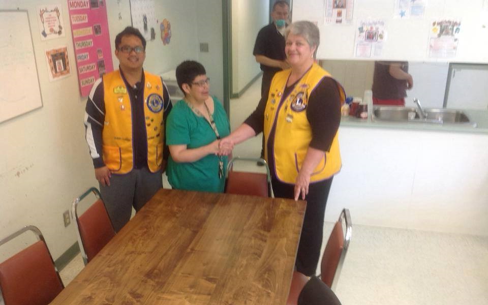 Three individuals in yellow vests stand together around a wooden table in a communal space, smiling and engaging with each other. The background features a kitchen area and various posters on the walls.